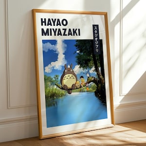 May include: A framed poster featuring Hayao Miyazaki's artwork. The image depicts a scene with a large, furry creature and other characters near a body of water. The poster has a wooden frame and is leaning against a white wall.