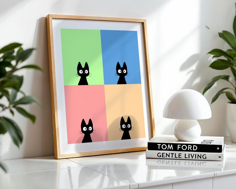 May include: A framed art print featuring four black cat silhouettes against a colorful background of green, blue, pink, and yellow. The artwork is displayed on a white surface next to a white mushroom-shaped lamp and two books.