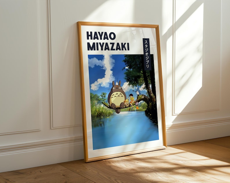 May include: A framed poster featuring Hayao Miyazaki's artwork. The image depicts a scene with a large, furry creature and other characters near a body of water. The poster has a wooden frame and is leaning against a white wall.