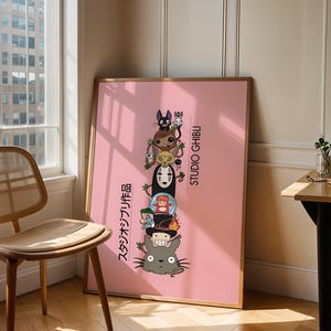 Puede incluir: Un p&oacute;ster rosa enmarcado con una pila de personajes de Studio Ghibli. Los personajes incluyen a Totoro, Sin Rostro y otros. El p&oacute;ster tiene texto japon&eacute;s y las palabras "STUDIO GHIBLI" en blanco. El p&oacute;ster est&aacute; apoyado contra una pared cerca de una ventana.