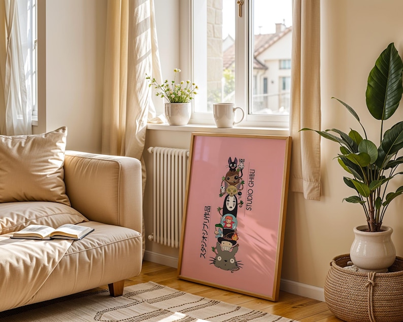 Puede incluir: Un p&oacute;ster rosa enmarcado con personajes de Studio Ghibli apilados, incluyendo a Totoro y Sin Rostro. El p&oacute;ster tiene texto japon&eacute;s y las palabras "STUDIO GHIBLI". El p&oacute;ster est&aacute; en una habitaci&oacute;n con un sof&aacute; beige y una planta.