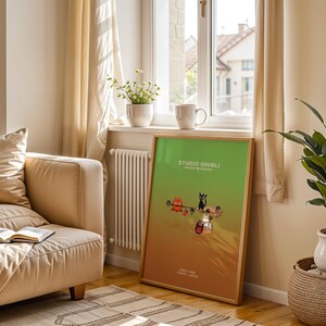 May include: A framed Studio Ghibli poster with a green and orange gradient background. The poster features cartoon characters and the text "STUDIO GHIBLI" and "SINCE 1985 TOKYO - JAPAN". The poster is leaning against a wall near a window.