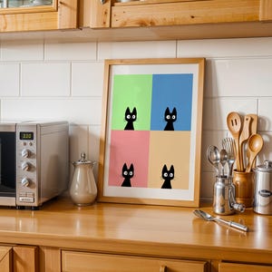 May include: A framed art print featuring four black cat silhouettes against a colorful background of green, blue, pink, and yellow. The artwork is displayed on a wooden countertop in a kitchen setting, alongside a microwave and kitchen utensils.