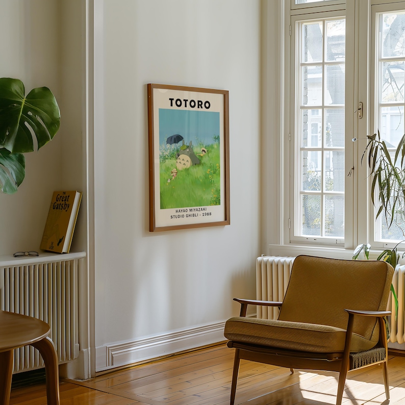 May include: A framed poster featuring the word "TOTORO" at the top, with an illustration of characters from the animated film. The poster is in a room with a wooden floor, a mustard-colored armchair, and a window.