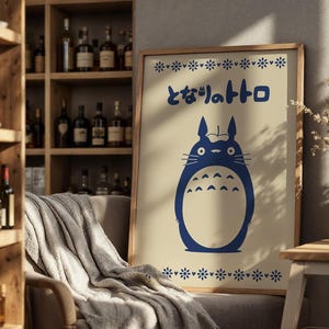 May include: A framed poster featuring a blue illustration of a cartoon character with the Japanese text "となりのトトロ". The poster has a beige background and a wooden frame. Decorative floral patterns are at the top and bottom of the poster.