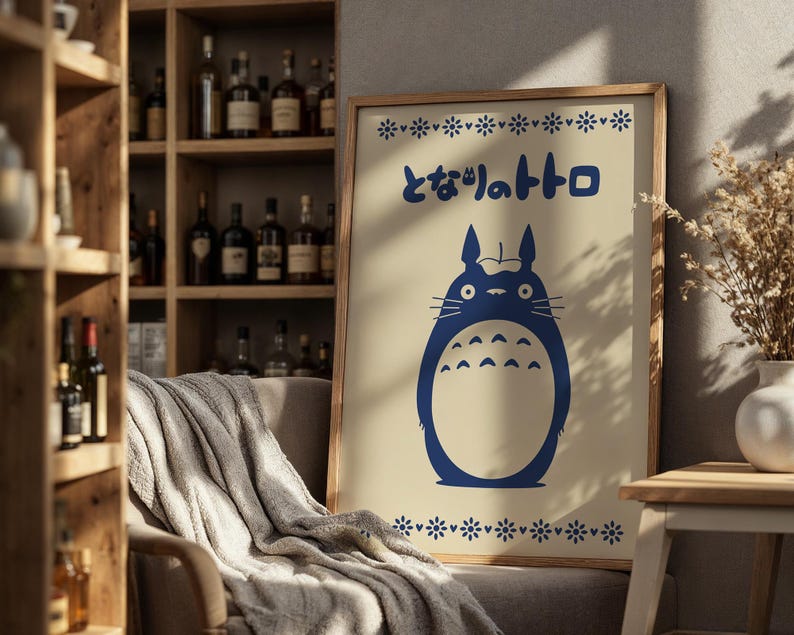 May include: A framed poster featuring a blue illustration of a cartoon character with the Japanese text "となりのトトロ". The poster has a beige background and a wooden frame. Decorative floral patterns are at the top and bottom of the poster.