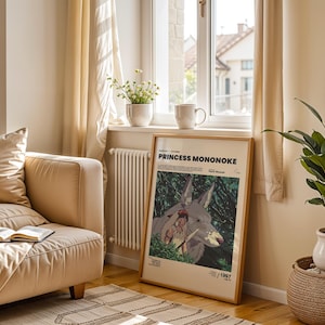 May include: A framed "Princess Mononoke" movie poster with a wooden frame, leaning against a wall. The poster features a wolf-like creature and a female figure in a forest setting. A beige sofa, a plant, and a window are also visible.