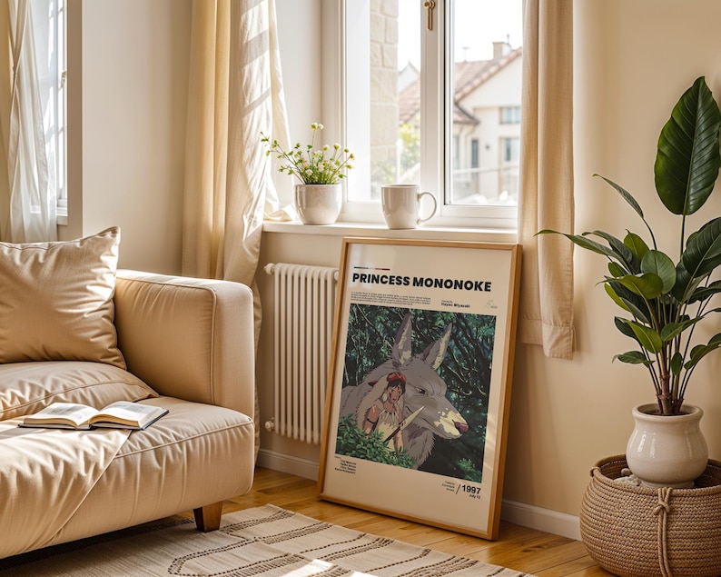 May include: A framed "Princess Mononoke" movie poster with a wooden frame, leaning against a wall. The poster features a wolf-like creature and a female figure in a forest setting. A beige sofa, a plant, and a window are also visible.