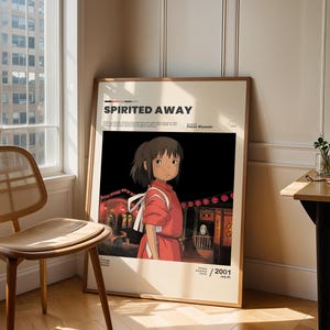 May include: A framed poster for the animated film "Spirited Away" featuring a character in a red dress. The poster has the film's title and the year 2001 printed on it. The poster is leaning against a wall next to a window and a wooden chair.