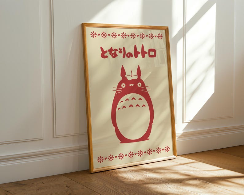 May include: A framed print featuring a red illustration of a Totoro character with Japanese text. The artwork is set against a cream background with decorative floral patterns at the top and bottom. The frame is a light brown color.