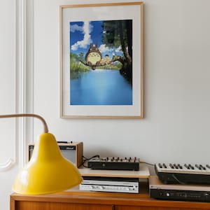 May include: A framed print of an animated scene featuring a large, furry creature and two children sitting on a tree branch over a body of water. The artwork is in a light wood frame, hanging on a white wall. A yellow lamp and stereo equipment are in the foreground.
