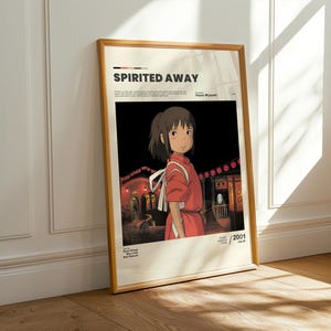 May include: Framed poster featuring the animated film "Spirited Away." The artwork depicts a young girl in a red outfit, set against a backdrop of traditional Japanese buildings illuminated at night. The poster has a wooden frame.