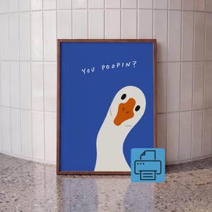Op de afbeelding: Een ingelijste kunstprint met een witte gans met een oranje snavel op een effen blauwe achtergrond. De tekst "YOU POOPIN?" staat in witte, handgeschreven letters in de linkerbovenhoek van het blauwe vlak boven de gans.