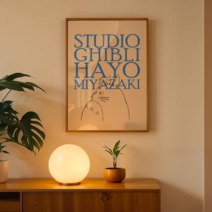 K&ouml;nnte beinhalten: Ein gerahmtes Poster mit blauem Text "STUDIO GHIBLI HAYO MIYAZAKI" und einer Katzenillustration. Eine leuchtende wei&szlig;e Kugellampe und eine kleine Topfpflanze stehen auf einem Holzschrank. Eine gro&szlig;e Topfpflanze befindet sich links.