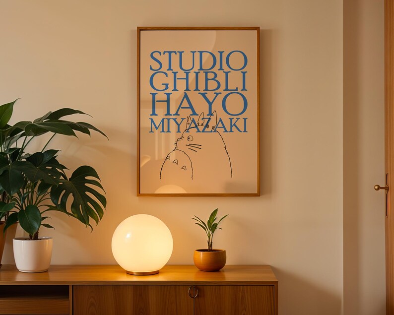 K&ouml;nnte beinhalten: Ein gerahmtes Poster mit blauem Text "STUDIO GHIBLI HAYO MIYAZAKI" und einer Katzenillustration. Eine leuchtende wei&szlig;e Kugellampe und eine kleine Topfpflanze stehen auf einem Holzschrank. Eine gro&szlig;e Topfpflanze befindet sich links.