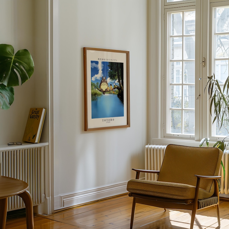 May include: A framed poster featuring a scene from the animated film "My Neighbor Totoro" hangs on a white wall. The poster has a brown frame and depicts a blue lake with characters from the film. A mid-century modern chair sits below the poster.