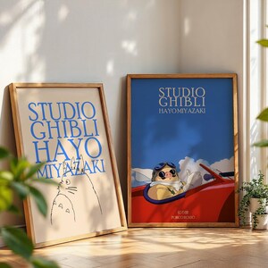 K&ouml;nnte beinhalten: Zwei gerahmte Poster mit Holzrahmen. Das linke Poster tr&auml;gt den blauen Text "STUDIO GHIBLI HAYO MIYAZAKI". Das rechte Poster zeigt eine Figur in einem roten Auto mit dem Text "STUDIO GHIBLI HAYO MIYAZAKI".