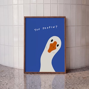 May include: A framed wall art print featuring a white goose with an orange beak against a solid blue background. The text "YOU POOPIN?" is printed in white, hand-drawn style lettering in the upper left corner of the blue field.
