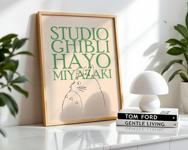 K&ouml;nnte beinhalten: Ein gerahmter Druck mit dem Text "STUDIO GHIBLI HAYO MIYAZAKI" in Gr&uuml;n und einer Strichzeichnung einer Figur. Eine wei&szlig;e pilzf&ouml;rmige Lampe steht auf zwei B&uuml;chern. Eine Topfpflanze ist im Hintergrund zu sehen.