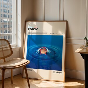 Op de afbeelding: Een ingelijste poster van de animatiefilm "Ponyo" van Hayao Miyazaki. De poster heeft een blauwe achtergrond met een illustratie van Ponyo in het water. De poster leunt tegen een muur naast een raam en een houten stoel.
