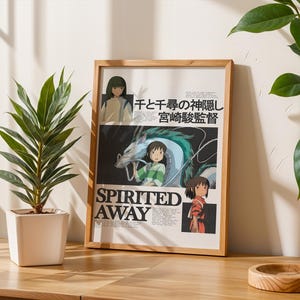 May include: A framed poster with artwork from the animated film "Spirited Away." The poster includes Japanese text and illustrations of characters and a dragon. The frame is light wood. A potted plant is to the left.