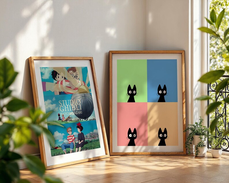 May include: Two framed art prints leaning against a wall. One features a Studio Ghibli movie poster with cartoon characters and text. The other has four black cat illustrations on a colorful background of green, blue, pink, and tan.