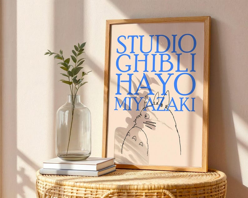 K&ouml;nnte beinhalten: Ein gerahmter Kunstdruck mit den Worten "STUDIO GHIBLI HAYO MIYAZAKI" in Blau und einer Cartoon-Illustration einer Figur aus einem Studio Ghibli-Film. Der Druck befindet sich in einem hellen Holzrahmen.