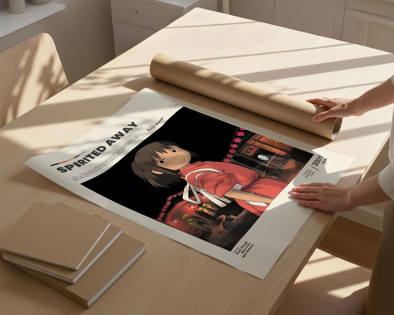 May include: A poster of the animated film "Spirited Away" featuring a character in a red outfit. The poster is on a light-colored table with a rolled-up poster and several square notebooks. The title "Spirited Away" is printed at the top.