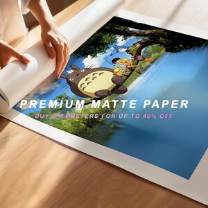 May include: A rolled-up poster featuring a scene with a large, gray cartoon character and two children near a body of water. The poster is being unrolled on a wooden surface. Text on the poster reads "PREMIUM MATTE PAPER".