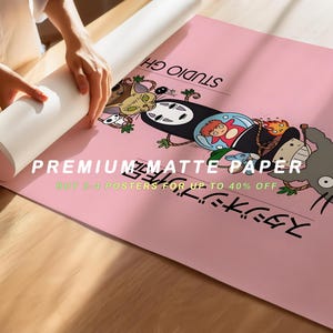 Puede incluir: Un p&oacute;ster rosa con personajes de una pel&iacute;cula de animaci&oacute;n japonesa. El p&oacute;ster tiene texto japon&eacute;s y las palabras "PREMIUM MATTE PAPER". Una persona est&aacute; desenrollando el p&oacute;ster sobre una superficie de madera.