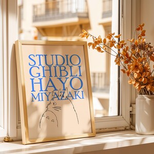 K&ouml;nnte beinhalten: Ein gerahmter Druck mit dem Text "STUDIO GHIBLI HAYO MIYAZAKI" in Blau, mit einer Schwarz-Wei&szlig;-Illustration von zwei Zeichentrickfiguren. Der Druck befindet sich in einem hellen Holzrahmen und wird auf einer Fensterbank ausgestellt.