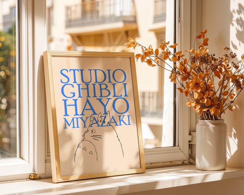 K&ouml;nnte beinhalten: Ein gerahmter Druck mit dem Text "STUDIO GHIBLI HAYO MIYAZAKI" in Blau, mit einer Schwarz-Wei&szlig;-Illustration von zwei Zeichentrickfiguren. Der Druck befindet sich in einem hellen Holzrahmen und wird auf einer Fensterbank ausgestellt.