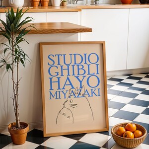K&ouml;nnte beinhalten: Ein gerahmtes Poster mit dem Text "STUDIO GHIBLI HAYO MIYAZAKI" in Blau, das eine Katzenillustration zeigt. Das Poster befindet sich in einem hellen Holzrahmen und lehnt an einer wei&szlig;en Wand. Eine Topfpflanze und ein Korb mit Orangen befinden sich im Vordergrund.