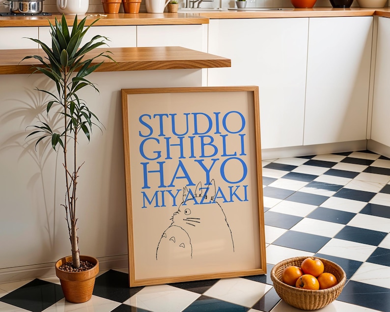 K&ouml;nnte beinhalten: Ein gerahmtes Poster mit dem Text "STUDIO GHIBLI HAYO MIYAZAKI" in Blau, das eine Katzenillustration zeigt. Das Poster befindet sich in einem hellen Holzrahmen und lehnt an einer wei&szlig;en Wand. Eine Topfpflanze und ein Korb mit Orangen befinden sich im Vordergrund.