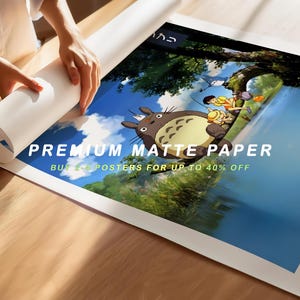 May include: A person unrolling a poster featuring an animated scene with a large, furry creature and other characters. The poster is printed on premium matte paper. Text on the poster reads "BUY 2+ POSTERS FOR UP TO 40% OFF".