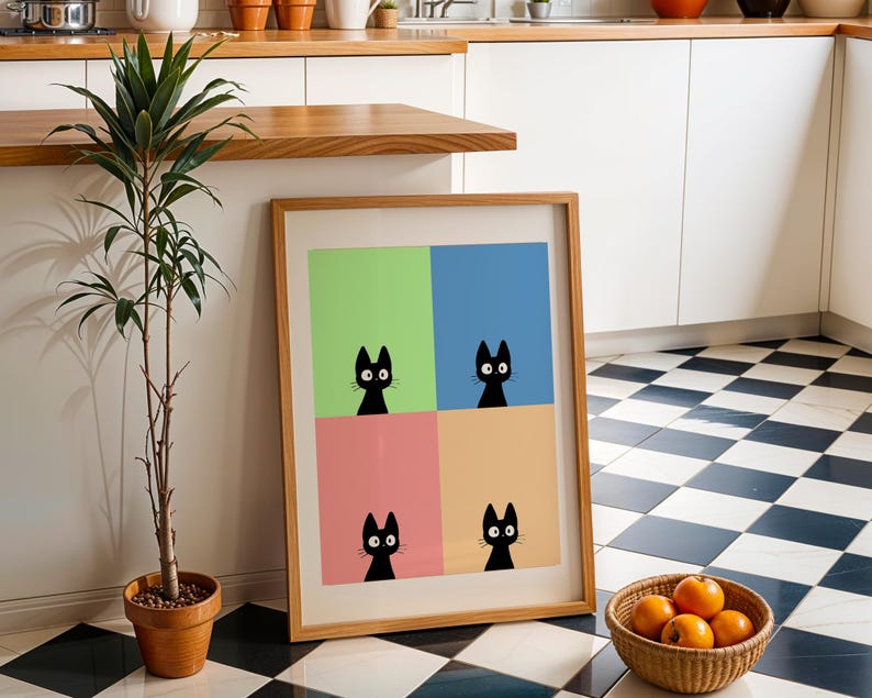 May include: A framed art print featuring four black cat illustrations against a colorful background of green, blue, pink, and orange squares. The artwork is displayed in a light wood frame, leaning against a wall next to a potted plant and a basket of oranges.