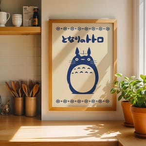 May include: Wooden framed print featuring a blue Totoro character with Japanese text. The artwork is displayed on a light-colored background with a floral border. The print is in a kitchen setting with wooden utensils and potted plants.
