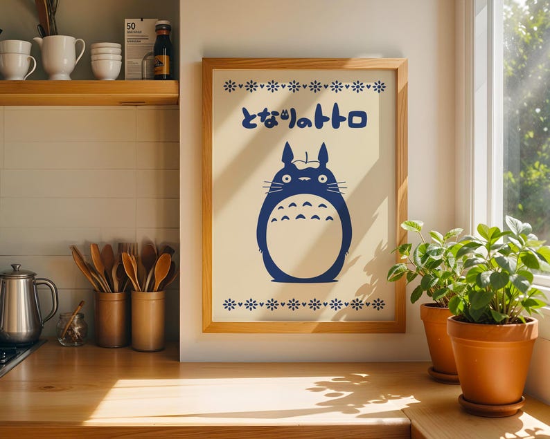 May include: Wooden framed print featuring a blue Totoro character with Japanese text. The artwork is displayed on a light-colored background with a floral border. The print is in a kitchen setting with wooden utensils and potted plants.