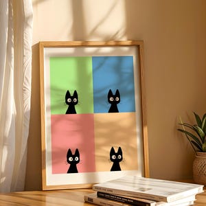 May include: A framed art print featuring four panels, each with a black cat silhouette against a different pastel background: green, blue, pink, and orange. The artwork is displayed on a wooden surface with books and a potted plant.