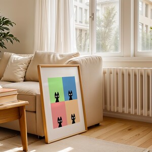 May include: A framed art print featuring four black cat illustrations against a colorful background of green, blue, pink, and orange. The artwork is displayed in a light wood frame, leaning against a beige sofa in a well-lit room with hardwood floors.