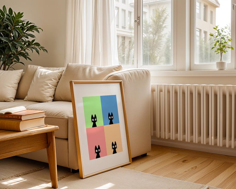 May include: A framed art print featuring four black cat illustrations against a colorful background of green, blue, pink, and orange. The artwork is displayed in a light wood frame, leaning against a beige sofa in a well-lit room with hardwood floors.