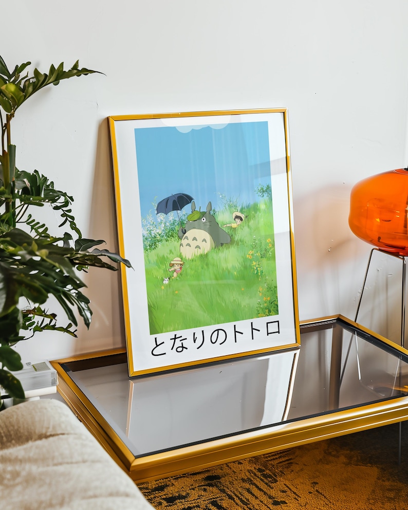 May include: Framed art print featuring a scene from the animated film, My Neighbor Totoro. The artwork depicts Totoro under an umbrella with children in a grassy field. The frame is gold, and the Japanese title is visible at the bottom.