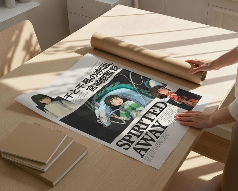May include: A poster with artwork from the animated film "Spirited Away" is displayed on a light wood table. The poster features Japanese text and character illustrations. A rolled-up brown paper tube and several brown notebooks are also on the table.