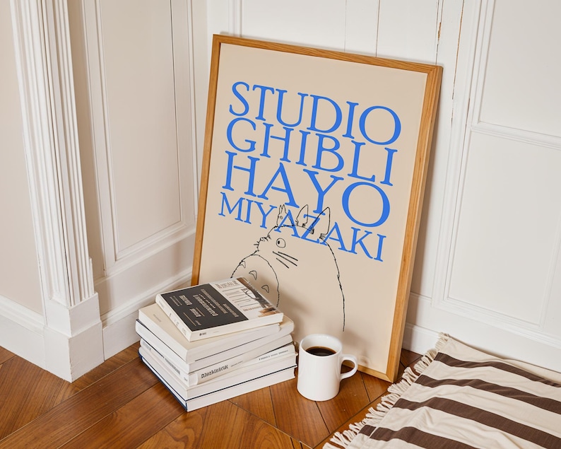 K&ouml;nnte beinhalten: Ein gerahmtes Poster mit blauem Text "STUDIO GHIBLI HAYO MIYAZAKI" und einer Cartoon-Illustration. Es lehnt an einer wei&szlig;en Wand, neben einem Stapel B&uuml;cher und einer wei&szlig;en Tasse mit Kaffee.