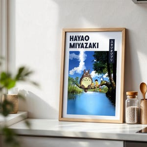 May include: Framed art print featuring Hayao Miyazaki's name and Japanese text. The artwork depicts a scene with a large, furry creature and two children sitting on a tree branch over a blue river. The background shows a blue sky and lush greenery.