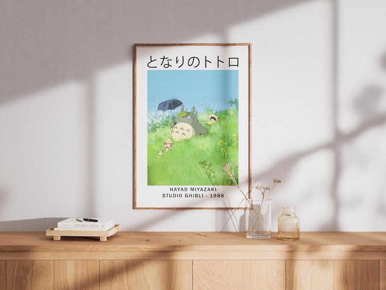 May include: A framed poster featuring a scene from the animated film "My Neighbor Totoro." The artwork depicts Totoro and other characters in a grassy field under a blue sky. Japanese text is at the top, with the film's title and studio information below.