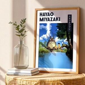 May include: Wooden framed poster featuring Hayao Miyazaki and Studio Ghibli. The artwork depicts Totoro sitting on a tree branch over a river, with a blue sky and clouds in the background. The poster is displayed on a wicker table.