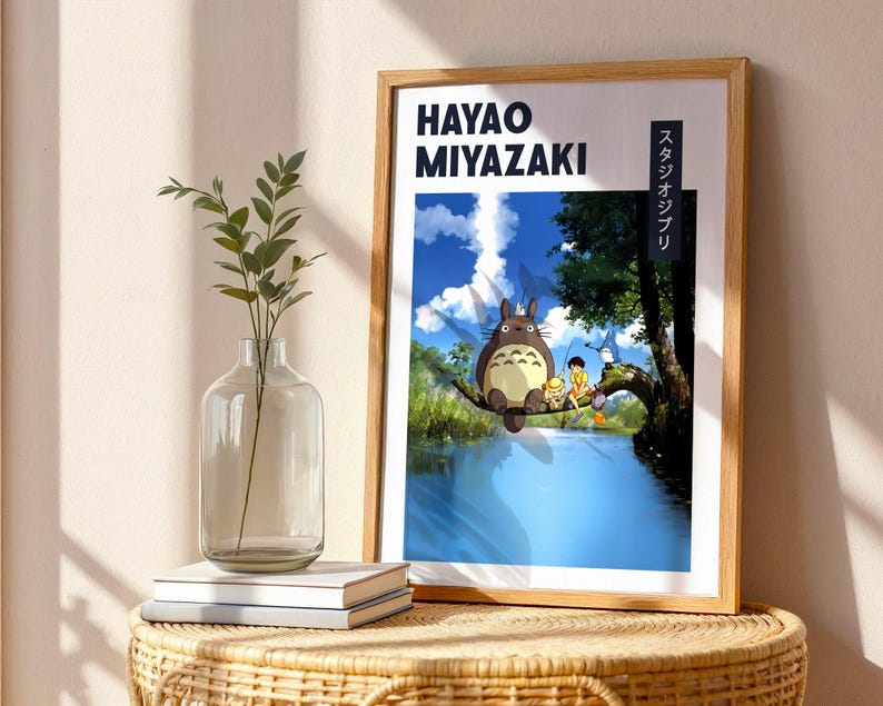 May include: Wooden framed poster featuring Hayao Miyazaki and Studio Ghibli. The artwork depicts Totoro sitting on a tree branch over a river, with a blue sky and clouds in the background. The poster is displayed on a wicker table.