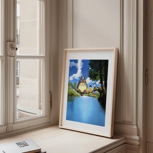 May include: A framed print featuring a scene with the character Totoro and children sitting on a tree branch over a blue river. The artwork has a light wood frame and is displayed on a windowsill next to a window.
