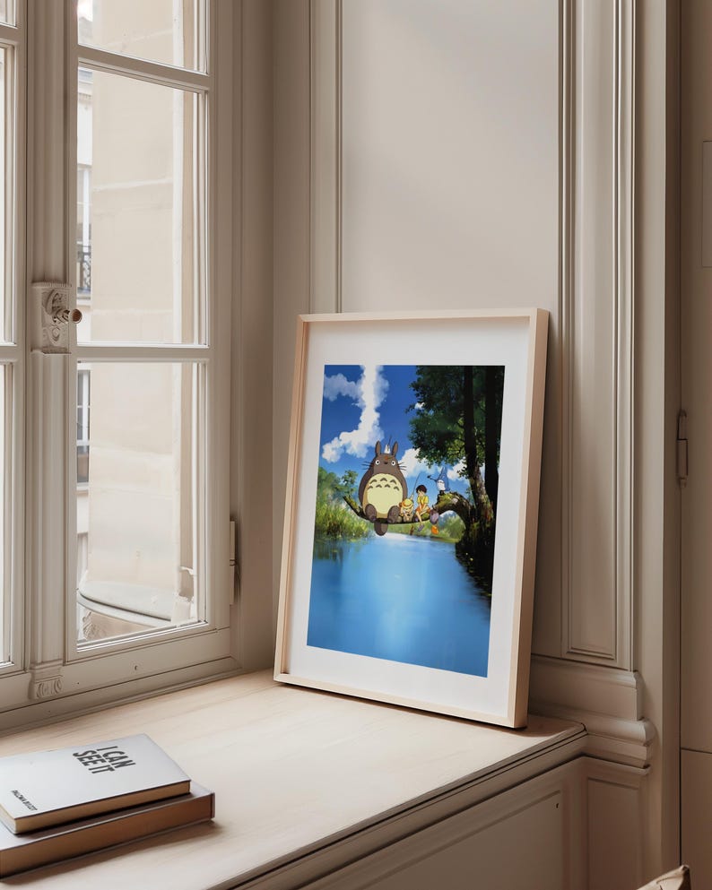 May include: A framed print featuring a scene with the character Totoro and children sitting on a tree branch over a blue river. The artwork has a light wood frame and is displayed on a windowsill next to a window.